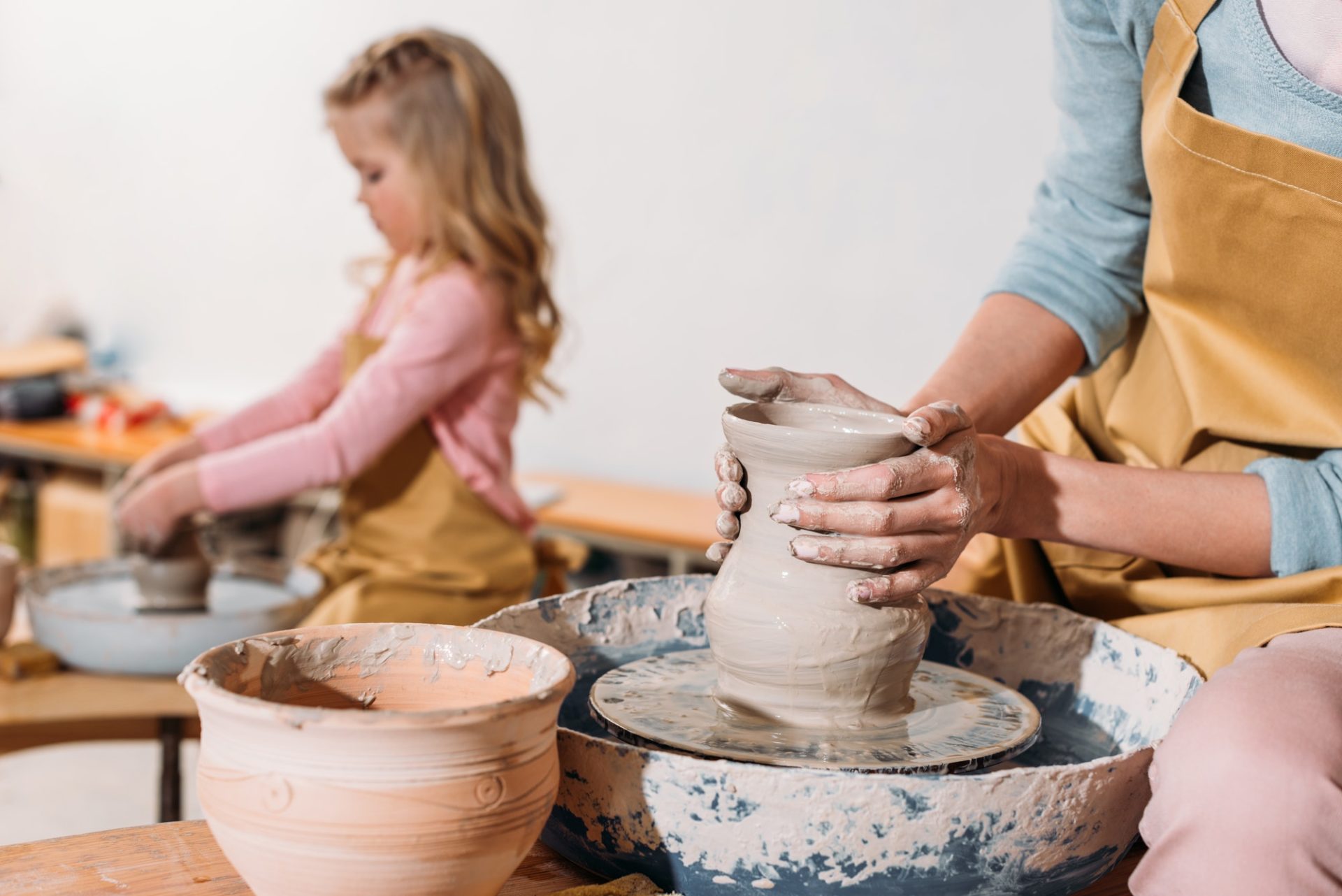 selective-focus-of-daughter-making-ceramic-pot-on-pottery-wheel-with-mother-on-foreground-e1656927048647.jpg